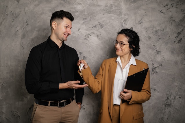 A person wearing a brown suit and holding a clipboard handing a set of keys to a person in a black button-down shirt